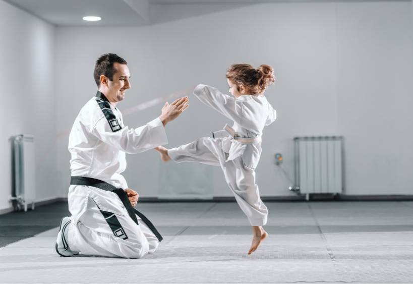 Entraîneur de taekwondo caucasien souriant en dobok, agenouillé et tenant la main levée pendant que la petite fille lui donne des coups de pied.