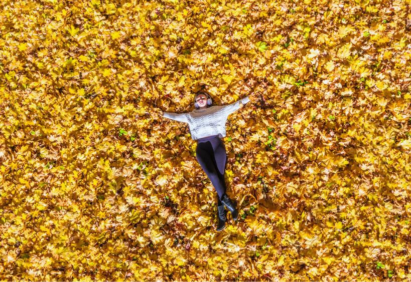 Jeune femme portant des écouteurs, allongée sur des feuilles d’érable d’automne dans un parc, écoutant de la musique et se détendant.