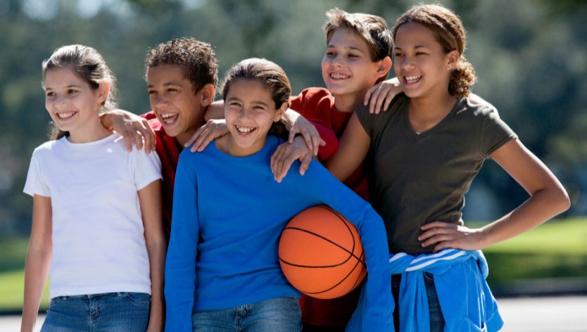 Cinq enfants souriants debout à l’extérieur, les bras autour des épaules les uns des autres, tenant un ballon de basketball, représentant l’esprit d’équipe et le plaisir dans un camp sportif pour jeun