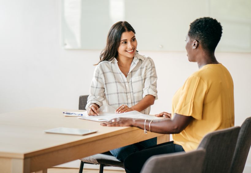 Deux femmes sont assises à une table dans un bureau lumineux, souriant et discutant de documents lors d’une réunion professionnelle.