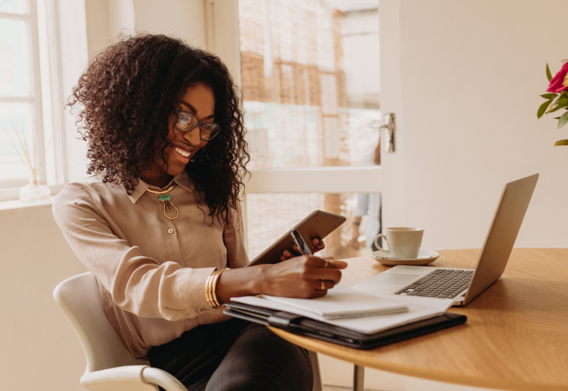 Femme d'affaires concentrée, assise à une table ronde avec tablette, stylo, ordinateur portable et café dans une pièce blanche