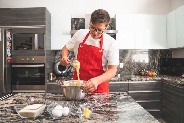 Adolescent portant un tablier rouge dans une cuisine moderne, fouettant des ingrédients dans un bol en acier inoxydable.