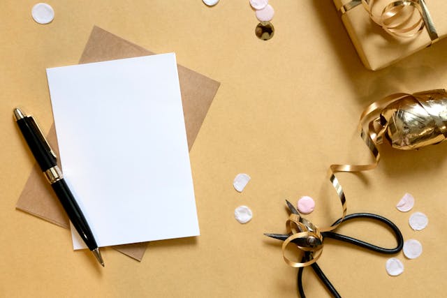 Carte vierge accompagnée d’une enveloppe brune, d’un stylo, de ciseaux et de décorations dorées
