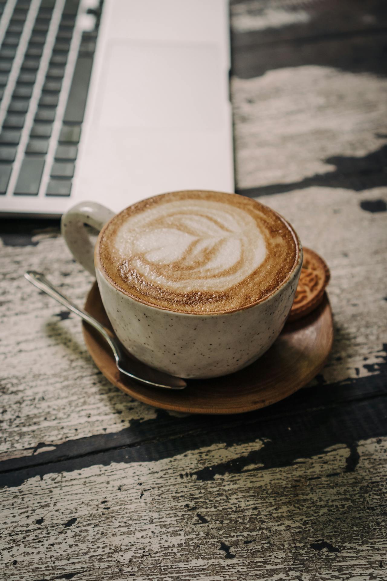 Gros plan d’une tasse de cappuccino et sa soucoupe sur une table rustique, à côté d’un ordinateur portable.