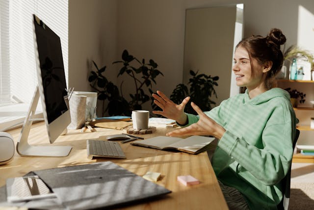 Jeune femme en sweat à capuche vert assise près d'une table, en train de discuter devant son ordinateur de bureau.