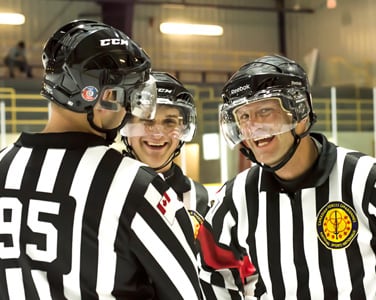 Three hockey referees in striped jerseys sharing a laugh on the ice rink.